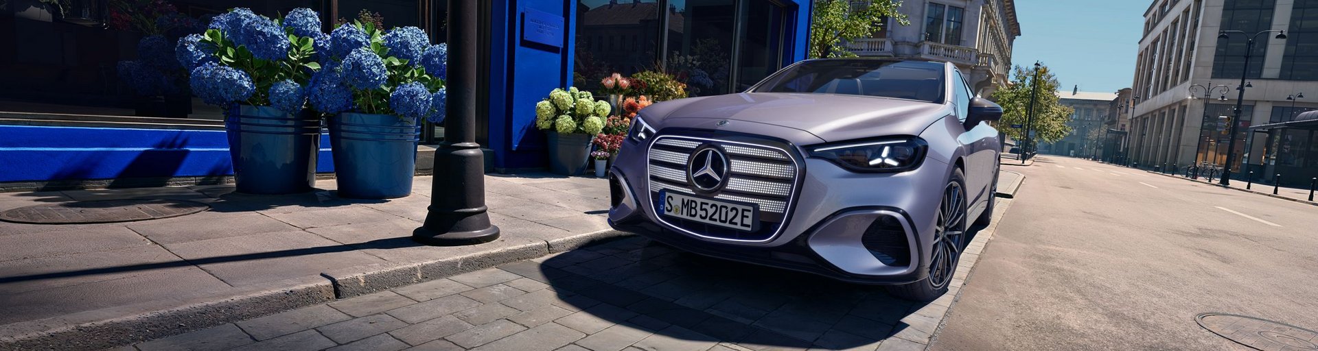 Mercedes-Benz C-Klasse elektrisch steht vor einem blauen Gebäude mit Blumendekoration auf einem sonnigen, leeren Straßenabschnitt in urbaner Umgebung