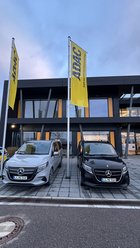 Zwei Mercedes-Benz Vans, ein silberner und ein schwarzer, stehen nebeneinander vor einem modernen Gebäude mit zwei gelben ADAC-Fahnen.