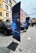 Zwei schwarze Autos stehen auf einem gepflasterten Platz vor mehrstöckigen Gebäuden, davor zwei aufrechte Werbefahnen mit dem Text 'Second Hand. First Class.' und Bildern von Autos.