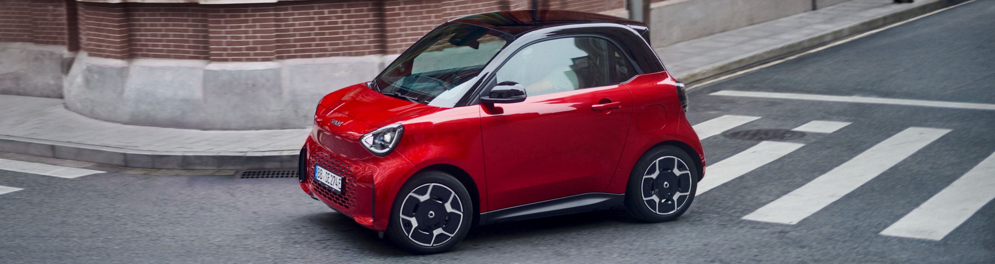 Roter smart #2 Kleinwagen mit schwarzem Dach fährt auf einer Kreuzung vor einem Backsteingebäude