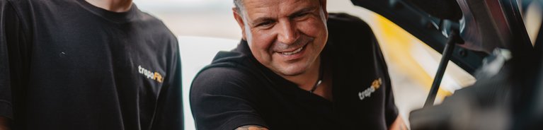 Zwei Männer in schwarzen T-Shirts mit gelbem Logo blicken unter die geöffnete Motorhaube eines Fahrzeugs