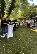 Drei Männer stehen an einem Stehtisch mit Getränken auf einer Wiese unter Bäumen, im Hintergrund mehrere Gruppen von Menschen bei einem Sommerfest mit Pavillon und Auto