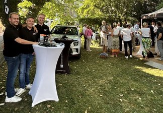 Drei Männer stehen an einem Stehtisch mit Getränken auf einer Wiese unter Bäumen, im Hintergrund mehrere Gruppen von Menschen bei einem Sommerfest mit Pavillon und Auto
