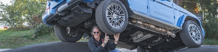 Mercedes-Benz G-Klasse steht schräg auf einer unebenen Asphaltfläche, während eine Person darunter kniet und das Fahrzeug mit den Händen zu stützen scheint