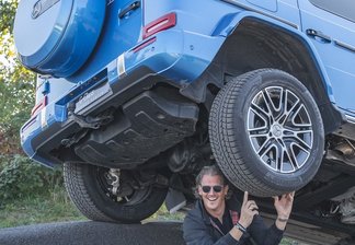 Mercedes-Benz G-Klasse steht schräg auf einer unebenen Asphaltfläche, während eine Person darunter kniet und das Fahrzeug mit den Händen zu stützen scheint