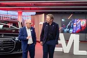 Zwei Männer im Anzug stehen vor einem schwarzen Mercedes-Benz und großen weißen Buchstaben 'EVL' in einem Autohaus mit LED-Bildschirm im Hintergrund