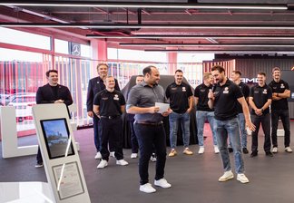 Gruppe von Personen in schwarzer Kleidung steht in einer modernen, offenen Halle mit großen Fenstern und sichtbaren Deckeninstallationen