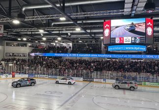 Eishockeyarena mit Zuschauern, drei Autos auf dem Eis und einer großen Anzeigetafel mit Werbung für BYD