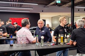 Mehrere Männer unterhalten sich an einem Stehtisch in einem Autohaus, auf dem Tisch stehen Getränke und kleine Vasen mit Blumen.