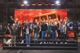 Gruppe von Menschen steht vor einer großen leuchtend orangen Leinwand mit der Silhouette eines Mercedes-Benz G-Klasse Fahrzeugs und hebt die Arme jubelnd in die Luft