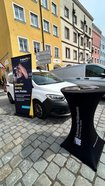 Stehtisch mit schwarzem Stretchbezug und Aufdruck 'Spartherm Vollkaskoschein' vor weißem Auto auf gepflastertem Platz mit gelbem und rosafarbenem Haus im Hintergrund